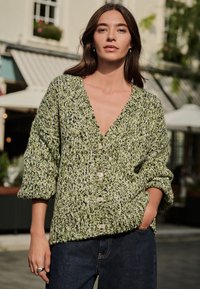 Grøn strikket cardigan med V-udskæring, store strukturerede masker og knaplukning. Har brede ærmer og ribbet kant.