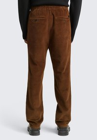 Bruine corduroy broeken met een ribbeltextuur, elastische tailleband en twee achterzakken met knoopsluitingen. Draag met zwarte schoenen.