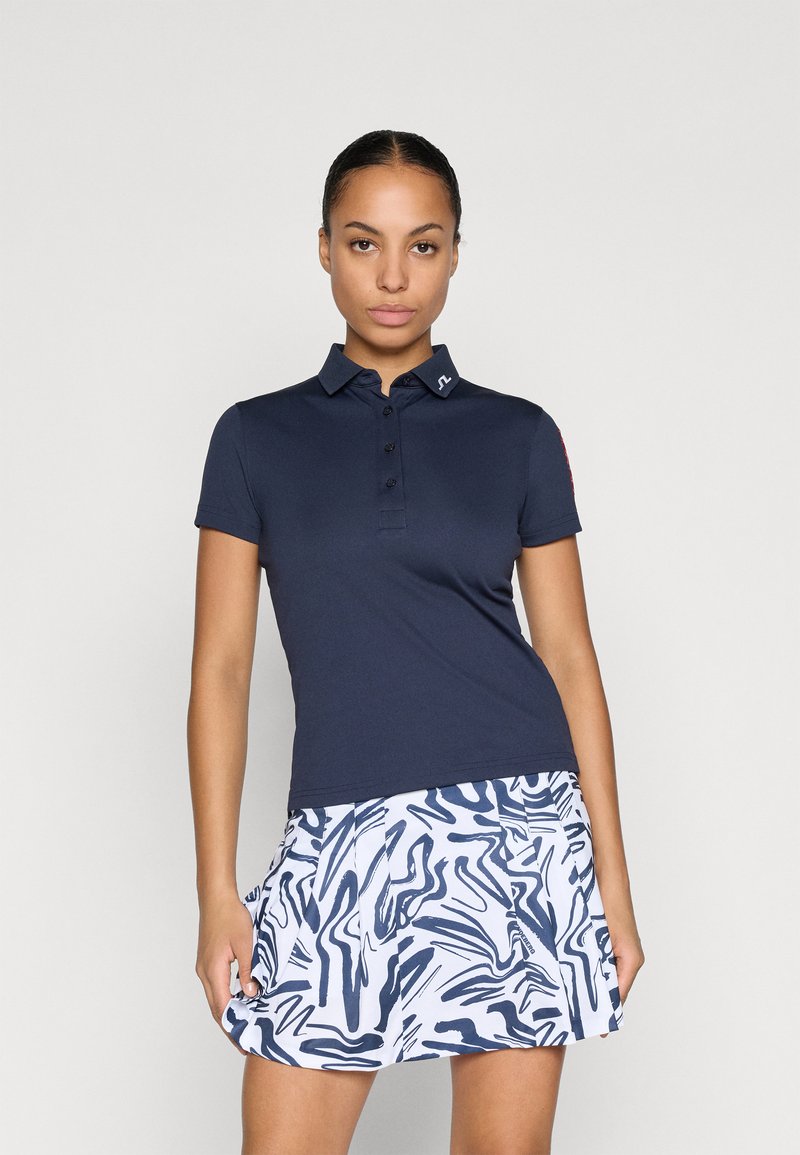 Jūras spēku polo krekls ar apkakli un pogu rindām, savienots ar baltu svārku, kuram ir abstrakts jūras spēku raksts. Abi izstrādājumi izgatavoti no viegla auduma.