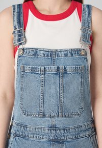 Denim overalls with a light wash, adjustable silver metal straps, and a front pocket. Paired with a white tank top featuring a red neckline.