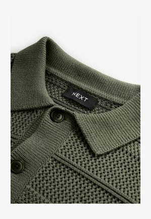 Olive roheline kootud polo särk, millel on tekstuuriga muster, kolm musta nuppu ja kaeluses silt "NEXT".