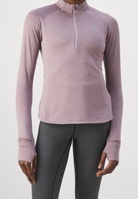 Person iført en lyserød langærmet sportstop med tommelhuller og sorte højresede leggings, stående med hænderne afslappet langs siden.