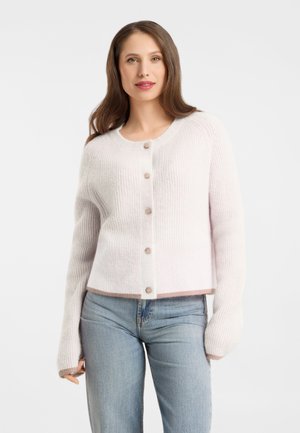 Junge Frau mit langen braunen Haaren, die eine weiße, geknöpfte Strickjacke und hellblaue Jeans trägt und vor einem einfarbigen Hintergrund steht.