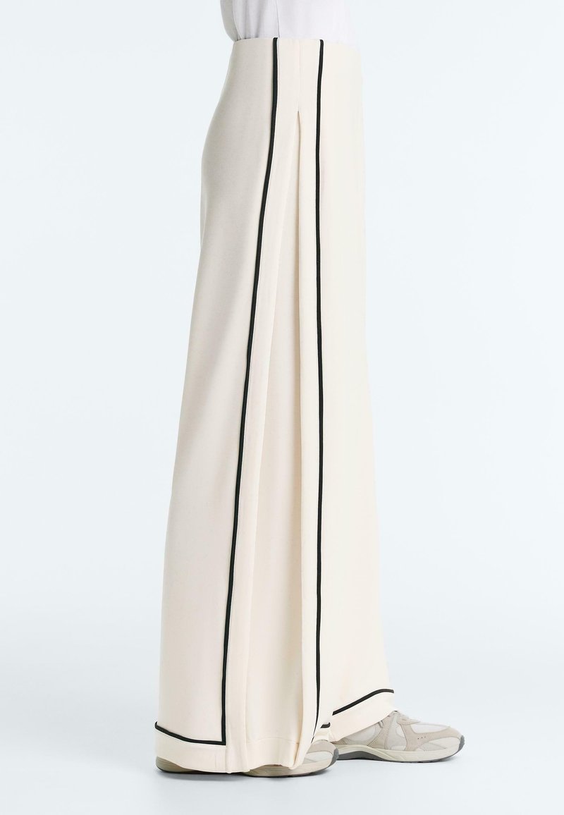 Pantaloni crem, cu talie înaltă și picioare largi, cu dungi verticale negre și tiv, purtați cu adidași albi și un top alb băgat în pantaloni.