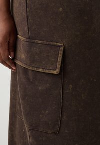 Fustă din țesătură maro, cu o suprafață texturată, având o mare buzunar cu o margine galbenă în contrast și detalii de cusătură vizibile.