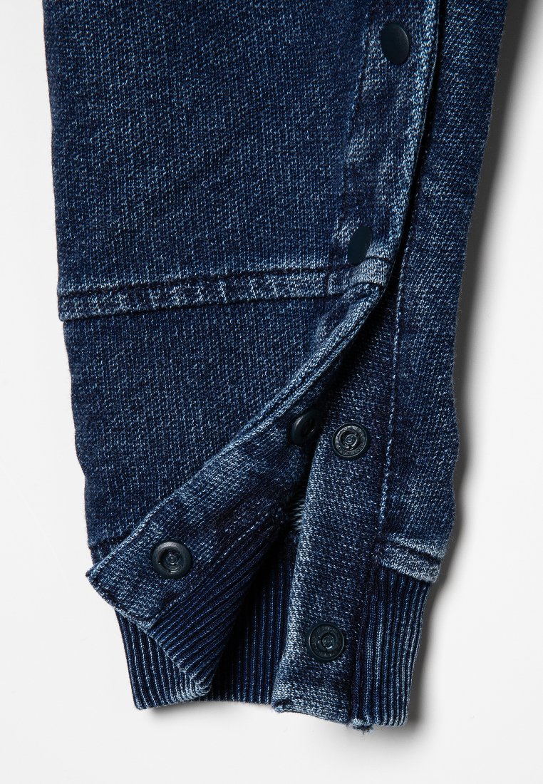 Dunkelblaue Jeanshose mit geknöpftem Saum, ausgestattet mit einer strukturierten Oberfläche, kontrastierender Nähte und fünf angenähten Knöpfen nahe dem Saum.