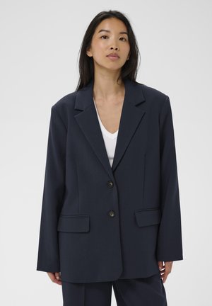 Młoda kobieta o długich czarnych włosach nosi ciemnogranatową, oversize'ową marynarkę na białej koszuli, stojąc na tle jasnego, jednolitego tła.