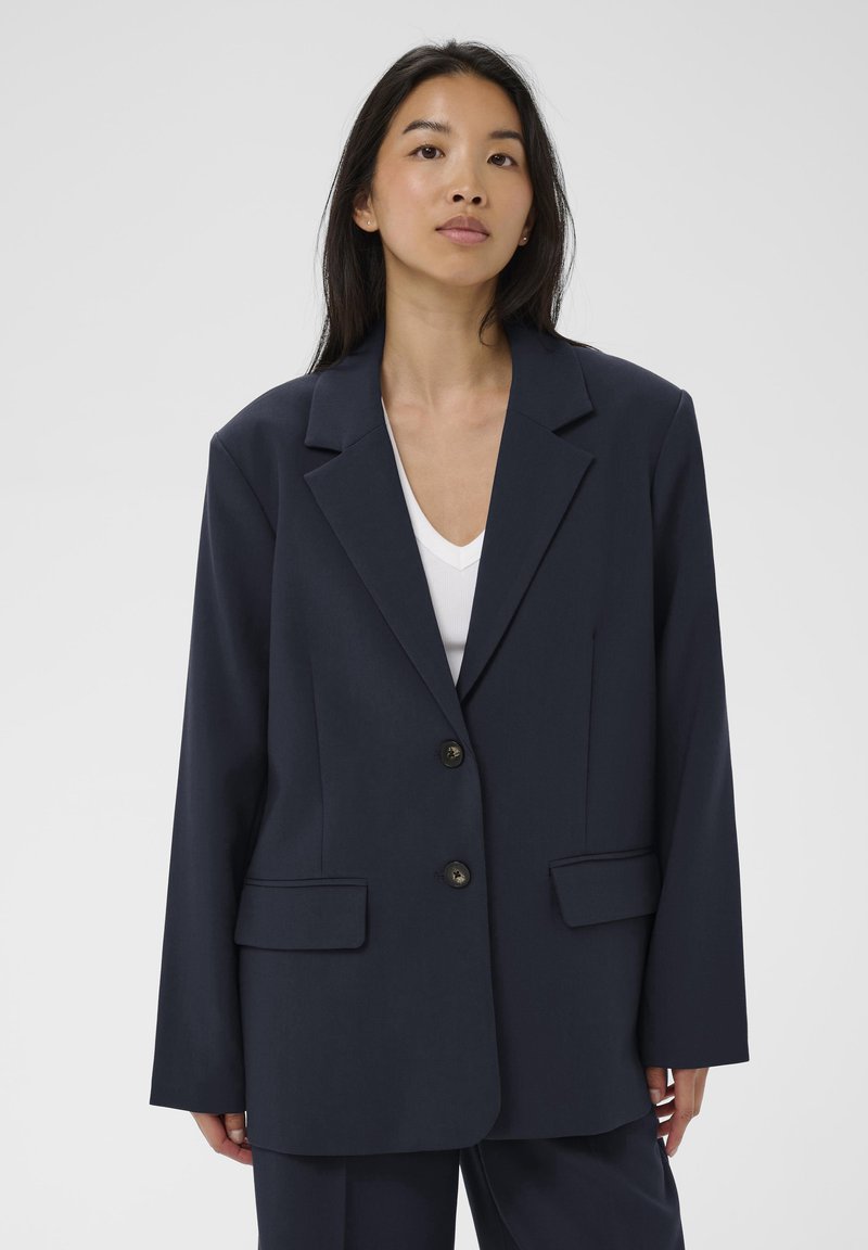 Joven mujer con cabello largo y negro lleva un blazer oversized de color azul marino oscuro sobre una camisa blanca, de pie contra un fondo claro y liso.