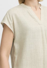 Blusa in lino beige chiaro con scollo a V e maniche corte. Il tessuto ha un motivo testurizzato, leggermente irregolare, che conferisce un aspetto informale.