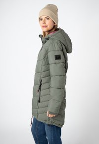 Vrouw in een beige gebreid mutsje, groene gewatteerde jas met capuchon en blauwe jeans, staande in profiel tegen een witte achtergrond.