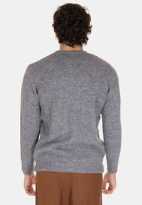 Uomo con capelli ricci scuri, che indossa un pullover grigio a maglia e pantaloni marroni, in piedi con la schiena rivolta verso l'osservatore su uno sfondo semplice.