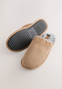Braune gestrickte Hausschuhe mit einem weichen grauen Innenfutter, einer Gummisohle und einem strukturierten Obermaterial, verziert mit einem kleinen Logo an der Seite.