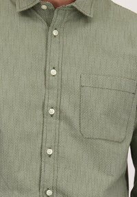 Knoopjeshemd in lichtgroen met een subtiel visgraatpatroon en een borstzak aan de rechterkant.