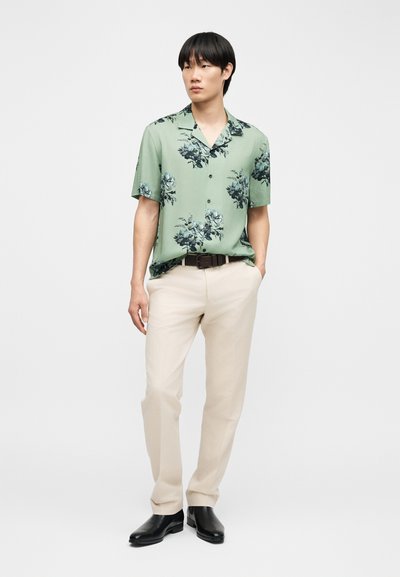 Άνδρας που φοράει ανοιχτοπράσινο κοντομάνικο πουκάμισο με λουλούδια, μπεζ παντελόνι, μαύρη ζώνη και μαύρα παπούτσια, στέκεται με το ένα χέρι στην τσέπη.