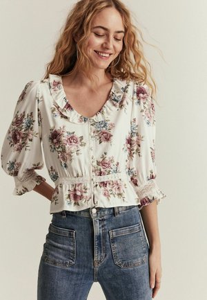 Blommig blus i krämfärgad med volangkrage och korta, puffiga ärmar. Har elastisk midja och spetsdetaljer. Matchad med höga blå jeans.