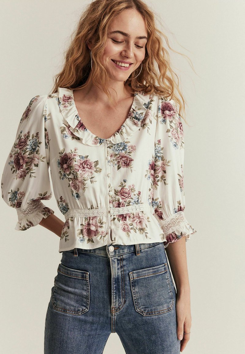 Blomstret bluse i creme med rynket halsudskæring og korte, puffede ærmer. Har elastisk talje og blondeaakcenter. Sættes sammen med højtaljede blå jeans.