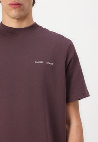 Lühikeste varukatega ümberkaela t-särk tumelilla kangast, millel on rinnal kaks valget tikandiga "SAMsøe" logo. Sile tekstuur, lihtne disain.