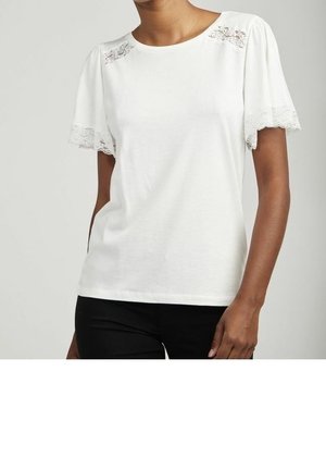 Kvinde iført hvid kortærmet top med blonder på ærmer og skuldre, kombineret med sorte bukser, stående foran en ensfarvet baggrund.