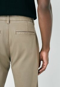 Rear view of a person wearing beige trousers and a black shirt, showing back pocket details and a relaxed arm hanging beside the body.