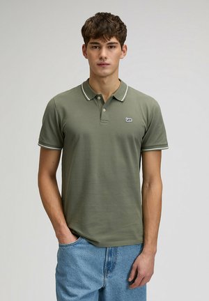 Młody mężczyzna ubrany w oliwkowo-zieloną koszulkę polo z białym wykończeniem i niebieskie dżinsy, stojący na tle jednolitego jasnego tła.