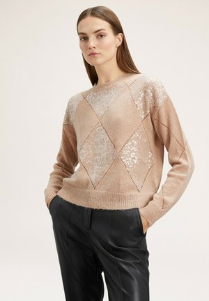 Beige gebreid trui met diamantpatronen, voorzien van paillettenaccenten op de schouders en een geribde onderrand. Gecombineerd met zwarte broeken.