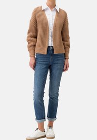 Kvinna i en beige chunky stickad cardigan över en vit skjorta med knappar, blå uppvikta jeans och vita sneakers med gummisulor.
