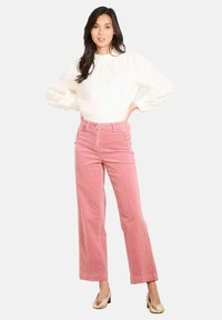 Žena stojí s rukami na bedrách, oblečená v bielej textúrovanej blúzke s dlhým rukávom, ružových širokých zamatových nohaviciach a zlatých lodiach na nízkom opätku.