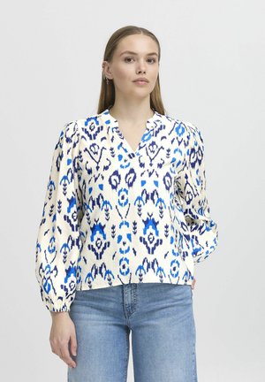 Blouse blanche avec un motif floral et en paisley bleu, décolleté en V, manches bouffantes et poignets élastiques, associée à un jean bleu clair.