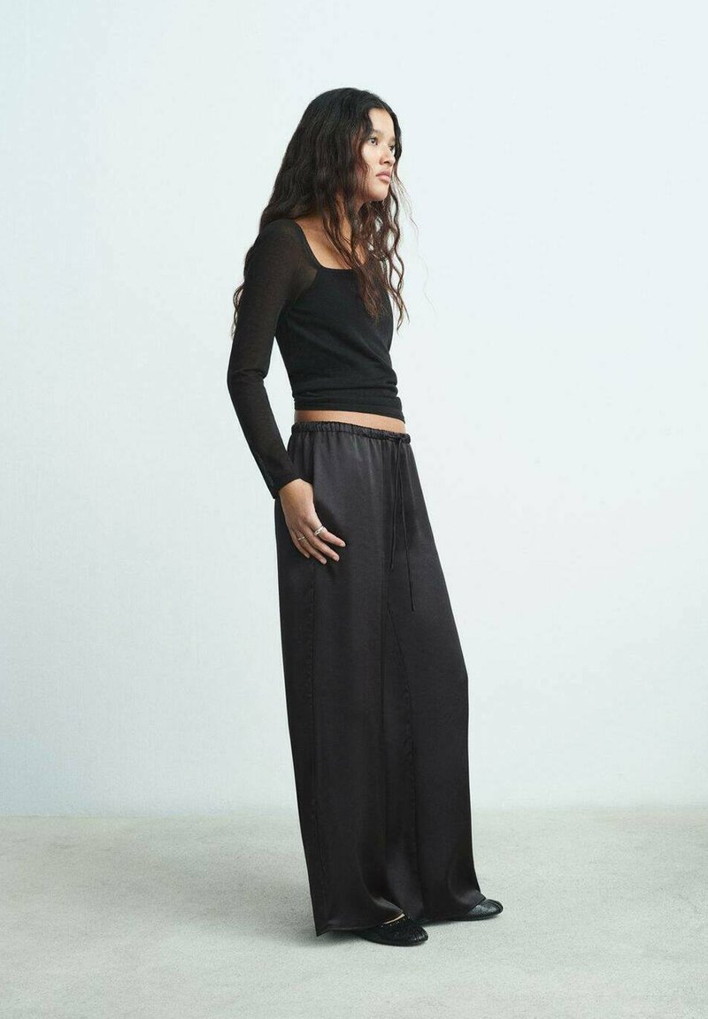 Schwarzes Langarmshirt kombiniert mit lockeren, hochgeschnittenen schwarzen Satin-Hosen mit elastischem Bund und Kordelzug.