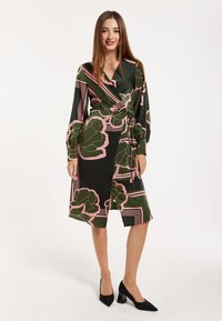 Wrap dress in dark green with large pink floral patterns and geometric lines. Long sleeves with tie detail at the waist. Black heels.