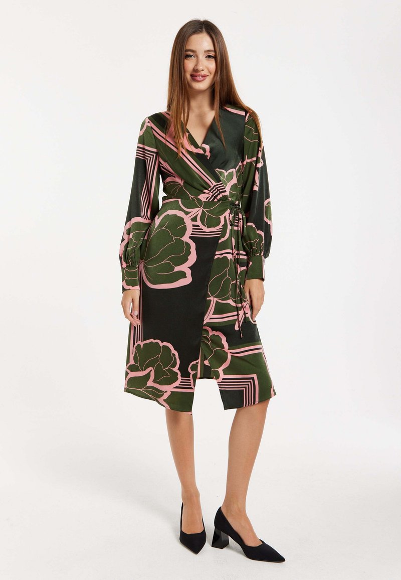 Wrap dress in dark green with large pink floral patterns and geometric lines. Long sleeves with tie detail at the waist. Black heels.
