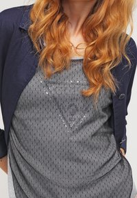 Veste bleu marine sur un débardeur gris avec un motif texturé et un design triangulaire en sequins. Tissu lisse et apparence superposée.