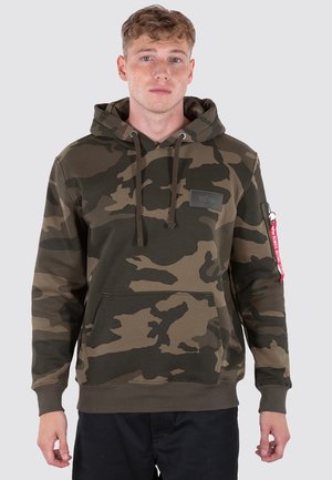 Young man wearing a green and brown camouflage hoodie with a front pocket and a red tag on the left sleeve, standing against plain background.