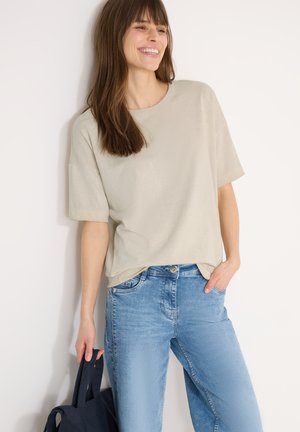 Donna sorridente appoggiata a un muro, indossa una camicia beige a maniche corte e jeans azzurri chiari, tenendo una borsa tote blu scuro al suo fianco.