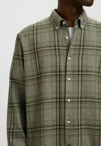 Zelena in siva karirana srajca iz mehkega materiala. Ima ovratnik z gumbi in bele gumbe spredaj.