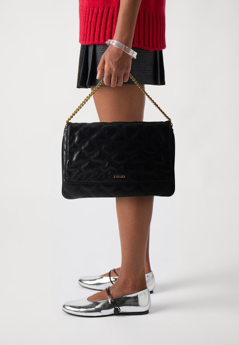 Personne portant un pull rouge et une jupe noire tient un sac à main noir matelassé avec une chaîne en or, portant des chaussures Mary Jane argentées.