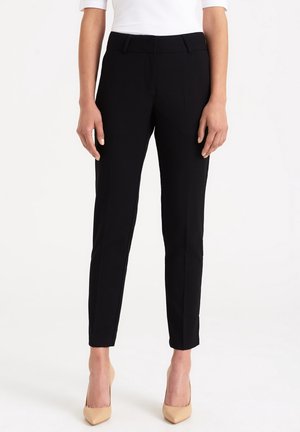 Vrouw draagt zwarte, nauwsluitende enkellange broek, witte top en beige puntige hakken, staand tegen een effen witte achtergrond.