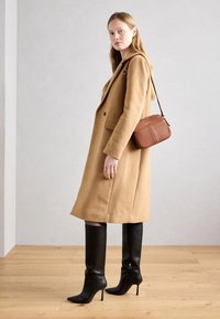 Velbloudí vlněný kabát se strukturovaným designem, černé kožené vysoké boty až ke stehnům a malá hnědá crossbody kabelka, umístěné na světlém pozadí.