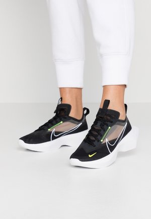 Personne portant des baskets Nike noires et blanches avec des accents vert néon et un pantalon de jogging blanc sur un sol blanc.