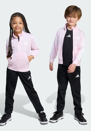 Dos niños con conjuntos deportivos a juego: una chaqueta de cremallera de color rosa claro con rayas blancas y pantalones negros con acentos blancos.