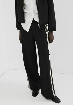 Persona vistiendo pantalones negros de pierna ancha con rayas blancas a los lados, chaqueta negra con cremallera, camiseta blanca y zapatos negros redondeados sobre fondo blanco.