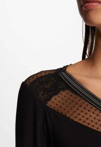 Zwarte jurk met een nettop met stippen en kantdetails, accentueert door een zilveren gestreepte halslijn. Glad stof op de mouwen.