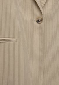 Tissu de blazer beige à fines rayures avec un seul bouton en écaille et une poche à raglan horizontale sur le côté gauche.