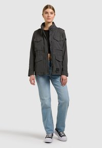 Jauna sieviete stāv, tērpusies tumši pelēkā darba jakā, melnā vēstures topā, gaiši zilās džinsa biksēs un melnbaltās sporta kurpēs, priekšā vienkārša fona.