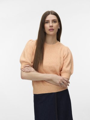 Femme aux longs cheveux raides portant un pull pêche à manches bouffantes et une jupe foncée, debout, les bras croisés, sur un fond uni.