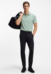 Lysegrøn polo med sort kant, sorte bukser og sorte sneakers. Holder en sort duffel-taske; har en logo på skjorten og bæltet.