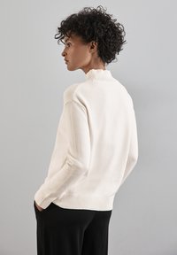 Lichtbeige trui met een hoge geribbelde kraag, lange mouwen en een geribbelde zoom. Zachte textuur, gedragen met zwarte broek.