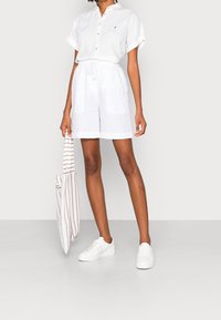 Vrouw in een witte korte mouwen blouse en witte shorts met trekkoord, met een gestreepte tote bag in de hand, staande met witte sneakers op een lichte achtergrond.