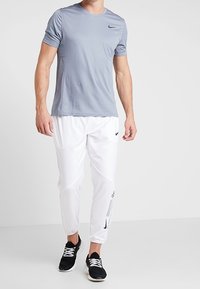 Muž v světlém šedém tričku Nike, bílých sportovních kalhotách Nike a černých sportovních botách, stojící na pozadí bílé plochy.