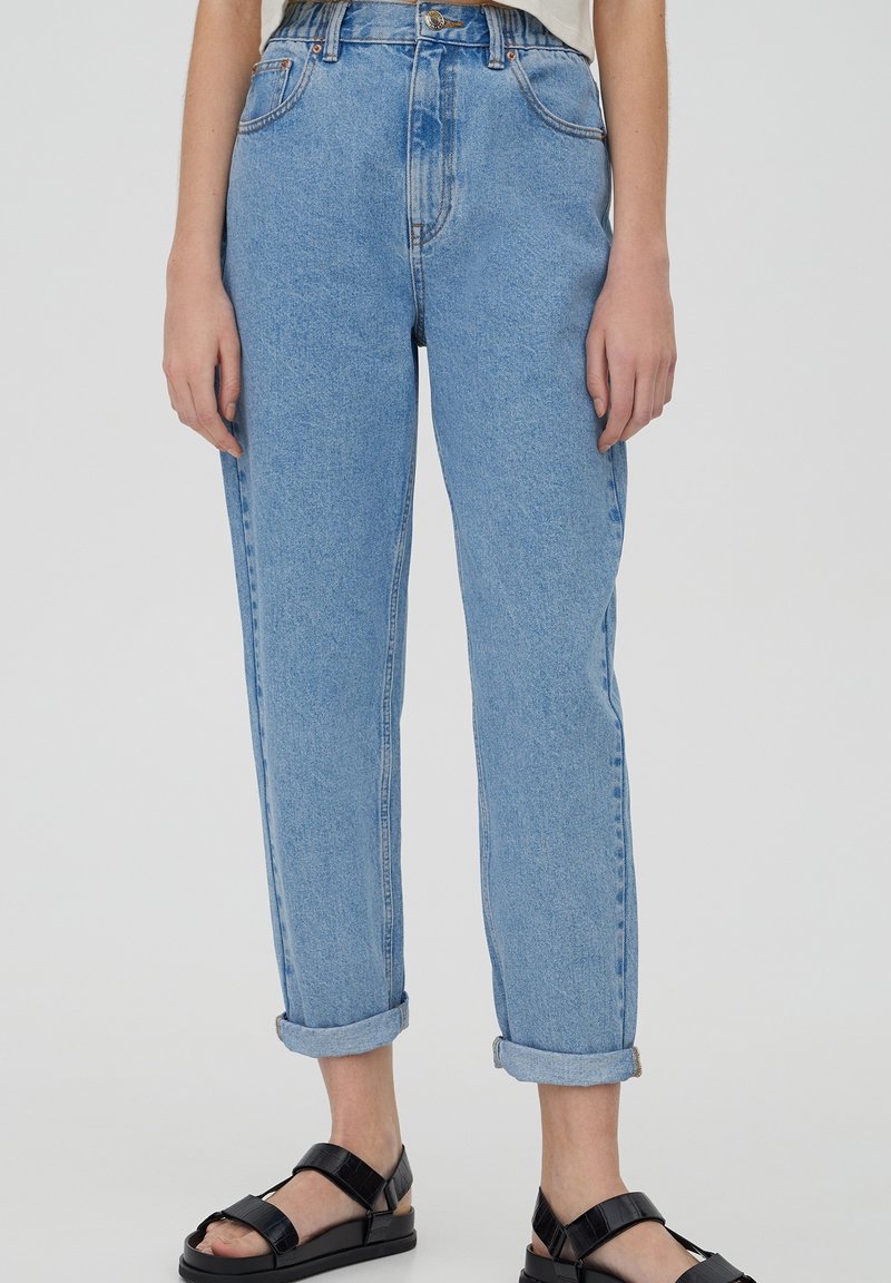 Jean taille haute bleu clair en denim, avec une coupe droite, des revers roulés et un design traditionnel à cinq poches. Sandales noires.